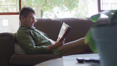 hombre caucásico sentado en el sofá, leyendo un libro en casa