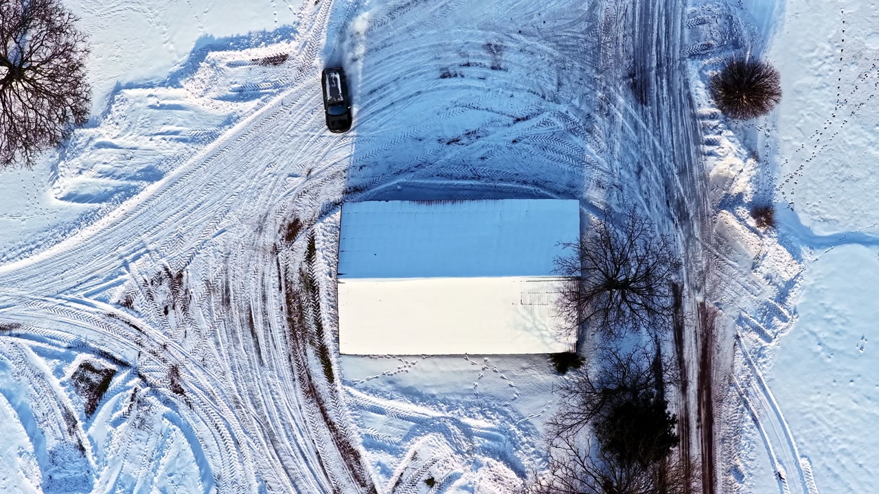 Aerial top-down captures car drifting on snow creating tire tracks around rural cottage amid trees snowy yard in Latvia winter sunrise