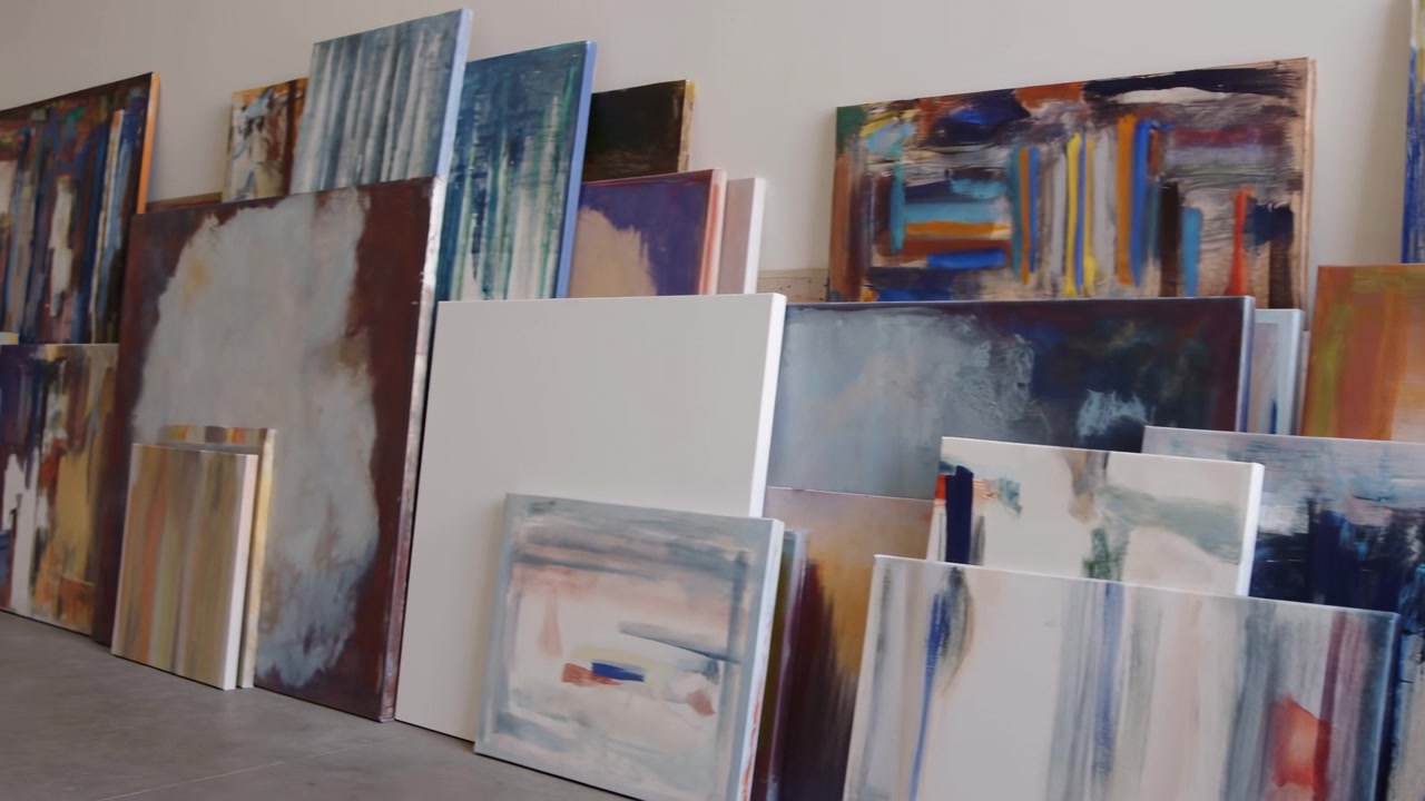 File di tele astratte senza cornice disposte in verticale e appoggiate a una parete dello studio bianco sporco, mostrando dimensioni varie, tecniche di pittura a strati, superfici materiche, neutri tenui, blu intenso e verde acqua.