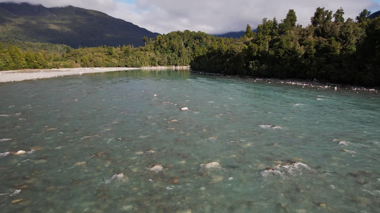 Hokitika River With Clear Shallow Water Flowing Over Stones With Dense Forest And Mountain In Background. West Coast, New Zealand. drone pullback shot