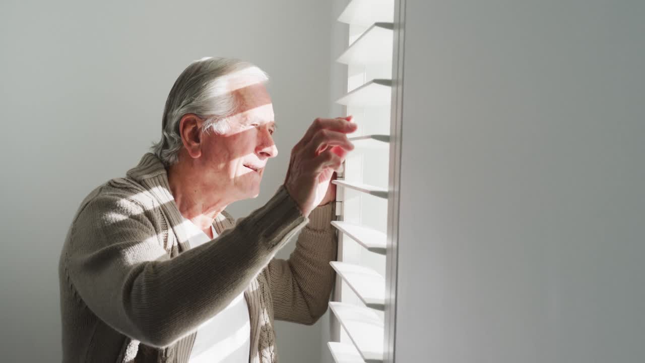Side view of retired senior man in cardigan sweater looking out through window blinds at home