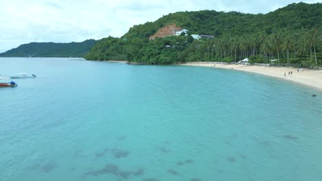 Aerial-shot-of-blue-sea,-boats,-sandy-beach,-green-forest,-and-hill