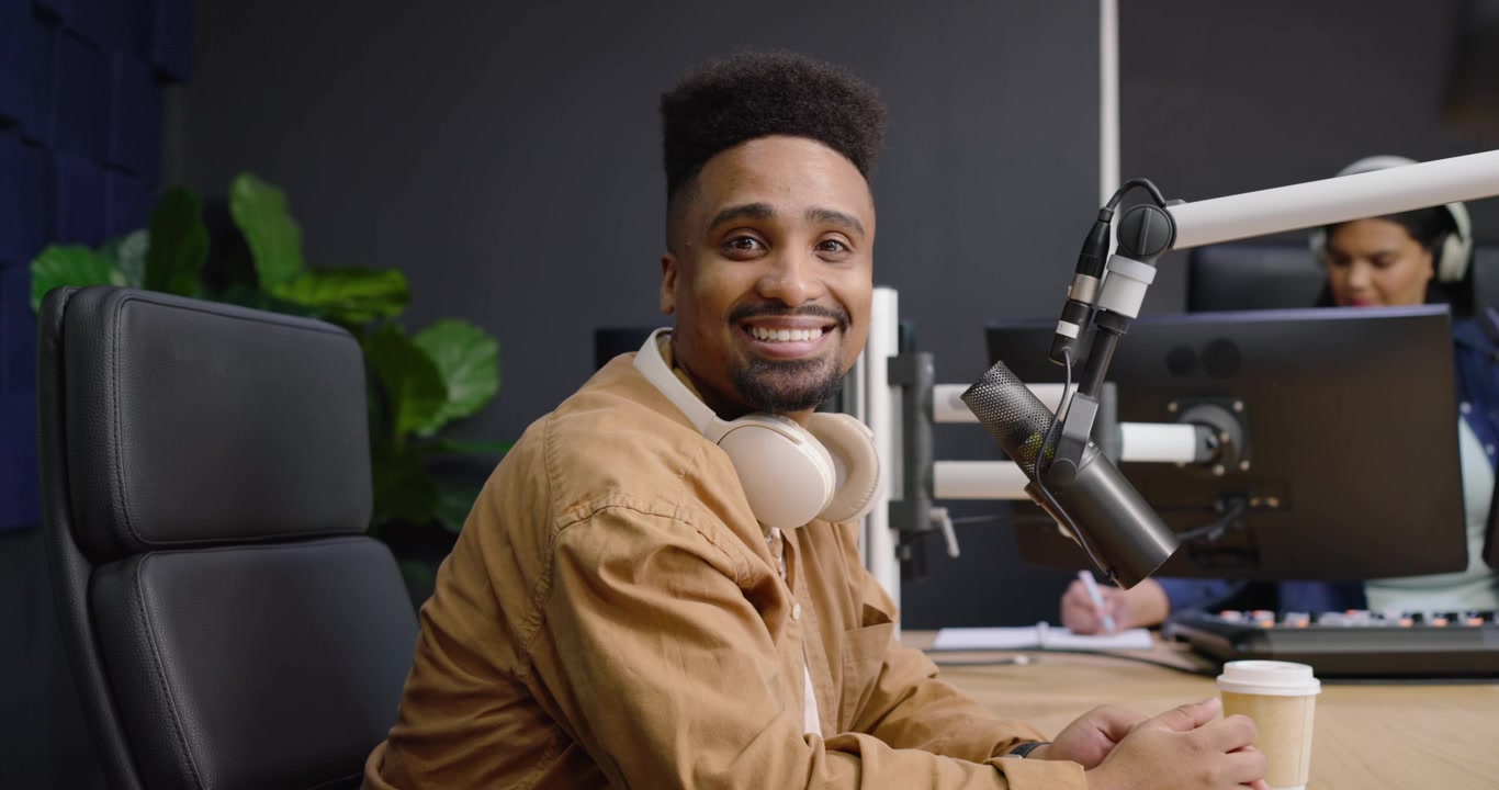 Uomo afroamericano con giacca color cachi che digita, sistema microfono e cuffie, sorridendo alla telecamera dello studio.