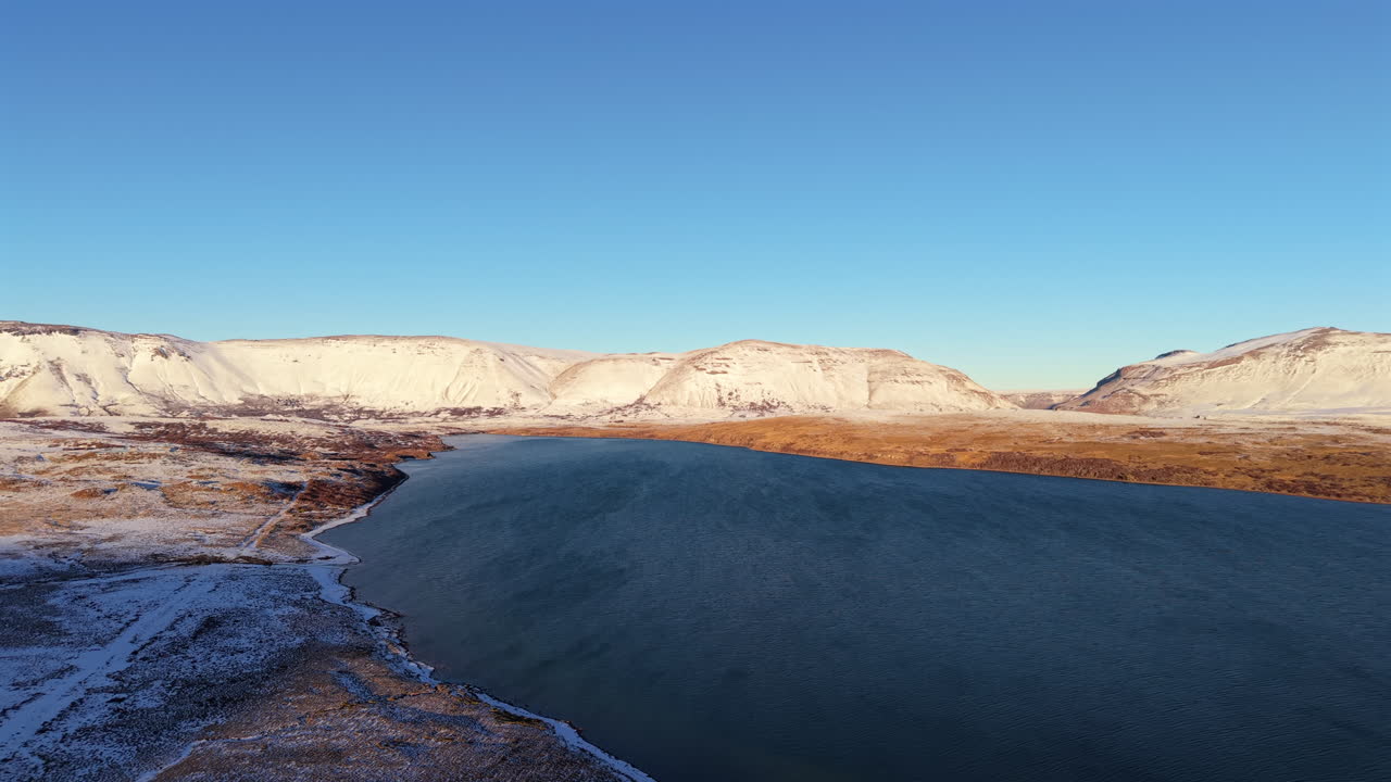 Aerial panning shot over Caviahue lake with snowy hills, rugged terrain, and clear blue sky in Patagonia, Argentina