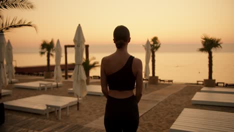 A-girl-with-tied-hair-in-a-black-sports-summer-uniform-goes-to-Golden-Beach-in-the-morning-to-do-yoga.-View-of-a-sunny-morning-beach-with-palm-trees-and-sun-loungers