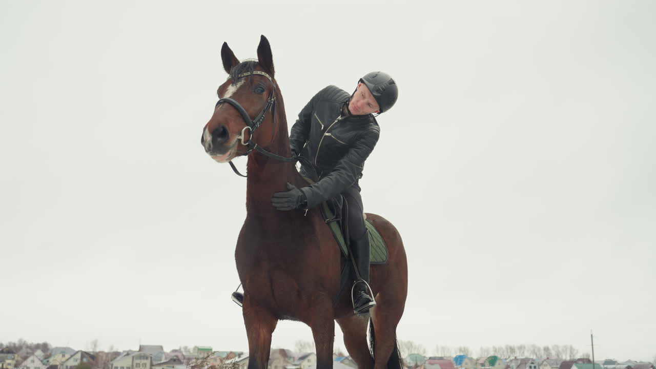 Cavaliere e cavallo in un campo innevato mostrano un legame in tandem durante una tranquilla lezione di equitazione, il cavaliere con il casco si china affettuosamente sulla sella, cavallo marrone con briglia, cielo coperto, villaggio distante, freddo.