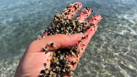 Hand-holding-and-playing-with-tiny-little-rocks-at-the-beach-with-turquoise-sea-water-and-waves-in-Manilva-Spain,-fun-sunny-summer-day,-4K-shot