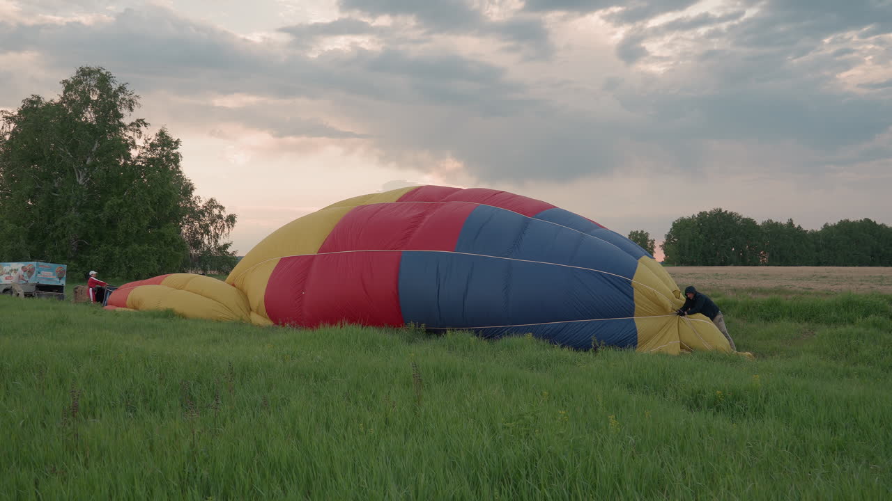 person in black hoodie and gloves pulling deflated multicolor hot air balloon envelope across lush green field under dramatic sunset sky with distant tree line for flight packing sequence