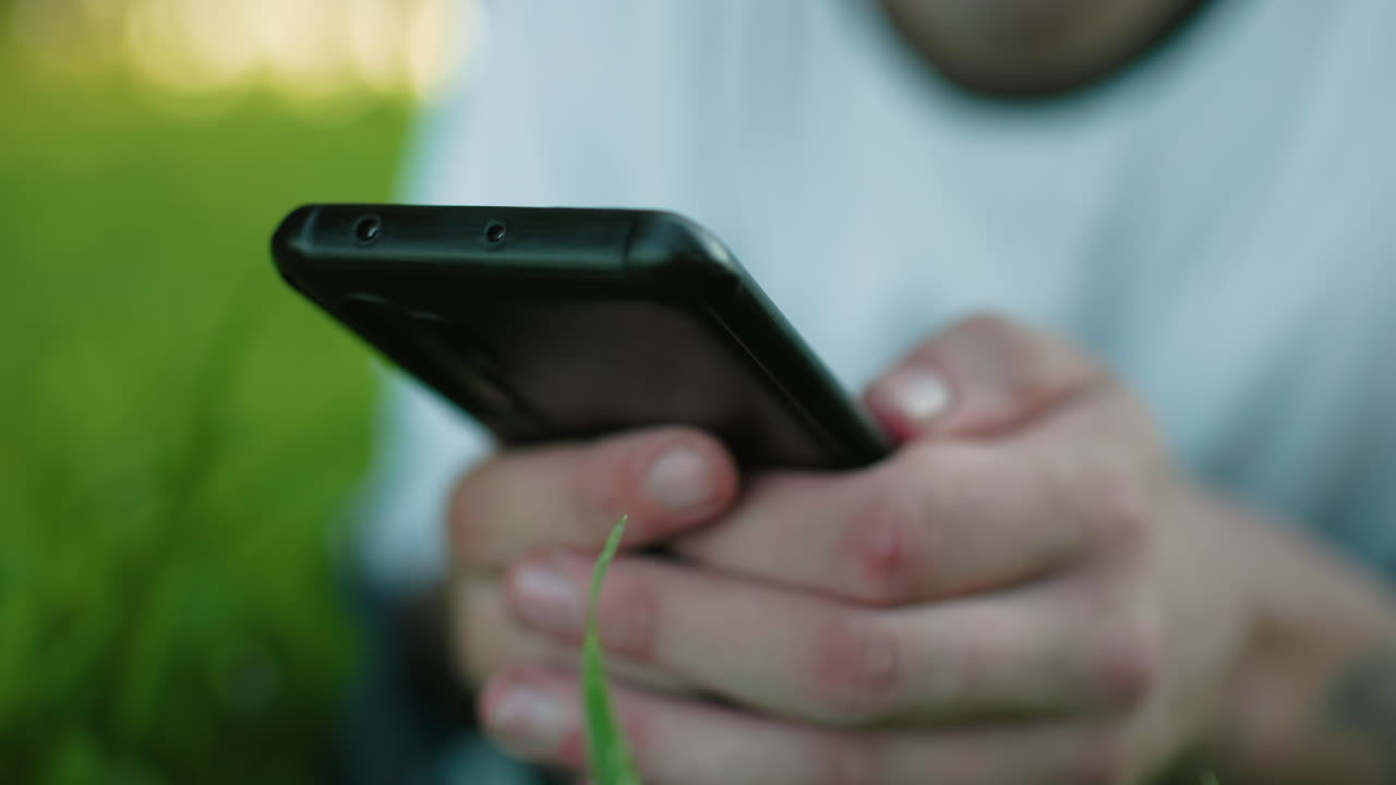 primer plano de un hombre tendido en un campo de hierba usando un teléfono inteligente, con enfoque en las manos sosteniendo el teléfono, el fondo natural borroso creando una atmósfera serena