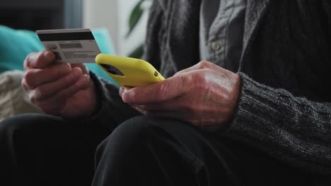 senior man enters credit card details into mobile phone for online shopping, sitting on sofa at home, close up