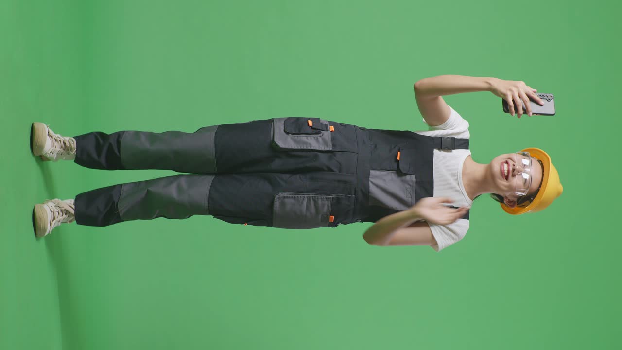 cuerpo lleno de trabajador mujer asiática con gafas de protección y casco de seguridad agitando la mano teniendo una llamada de video en el teléfono inteligente mientras está de pie en el estudio de fondo de la pantalla verde
