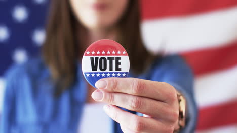 Kaukasische-Frau-Hält-Ein-&quot;Vote&quot;-Abzeichen,-Im-Hintergrund-Die-Amerikanische-Flagge