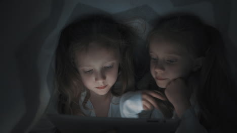 two little girls using a tablet covered by the bed sheet at night