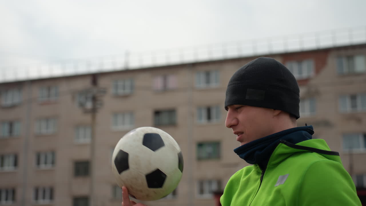 Крупный план мужчины, балансирующего футбольным мячом в квартире в городе, сосредоточенный спортсмен в повседневной одежде, отрабатывающий манипуляции с мячом на фоне городского пейзажа в помещении под облачным небом