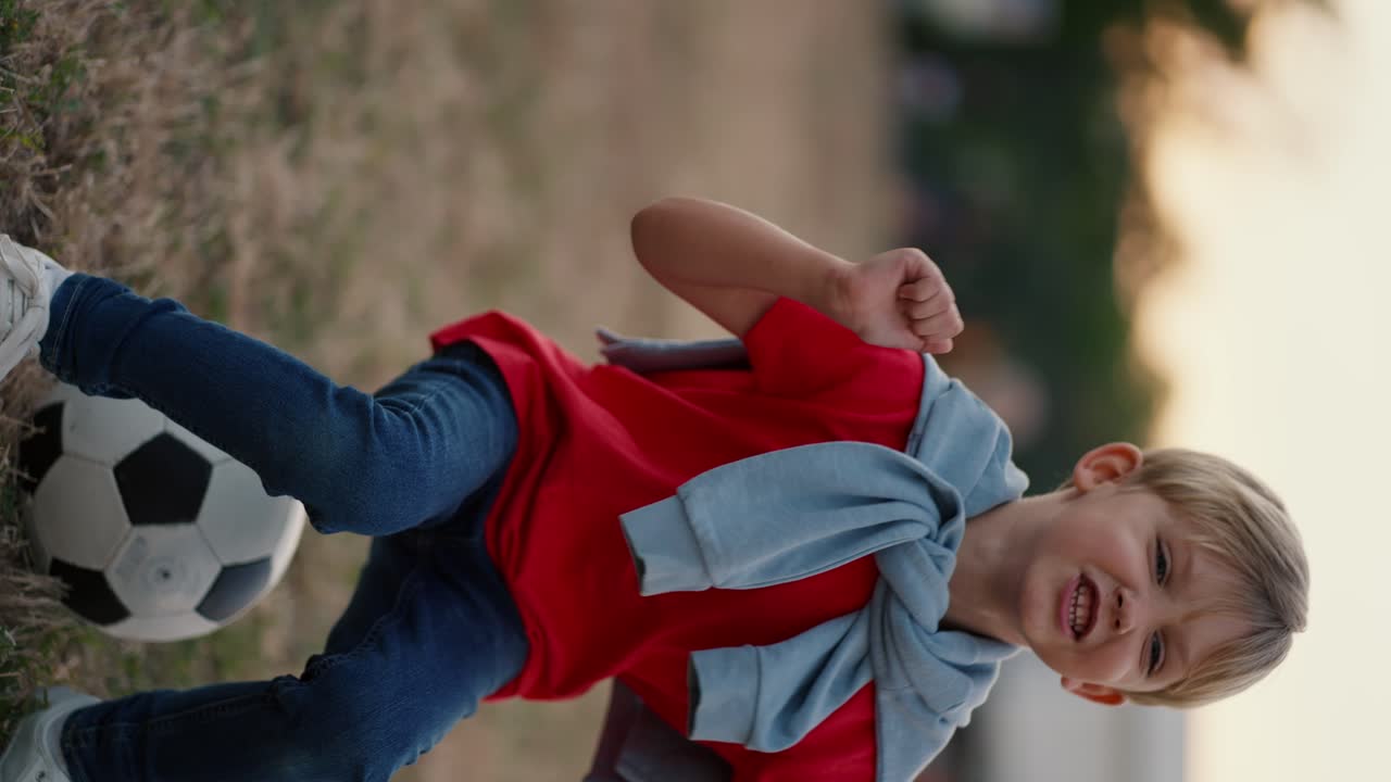 Vertical video A little blond boy with blue eyes in a red T-shirt and jeans jumps on a soccer ball on the playground in the park. Little boy having fun and sitting on a soccer ball on the soccer field in the park