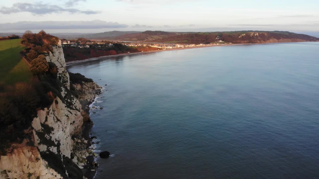 toma aérea de 4k sobre el mar y asombrosos acantilados que exponen la ciudad iluminada por el sol de beer england en el reino unido