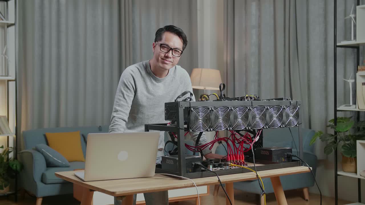 가상화폐 채굴을 위해 노트북과 채굴 기계를 사용하는 동안 카메라에 미소 짓는 안경을 쓴 아시아인