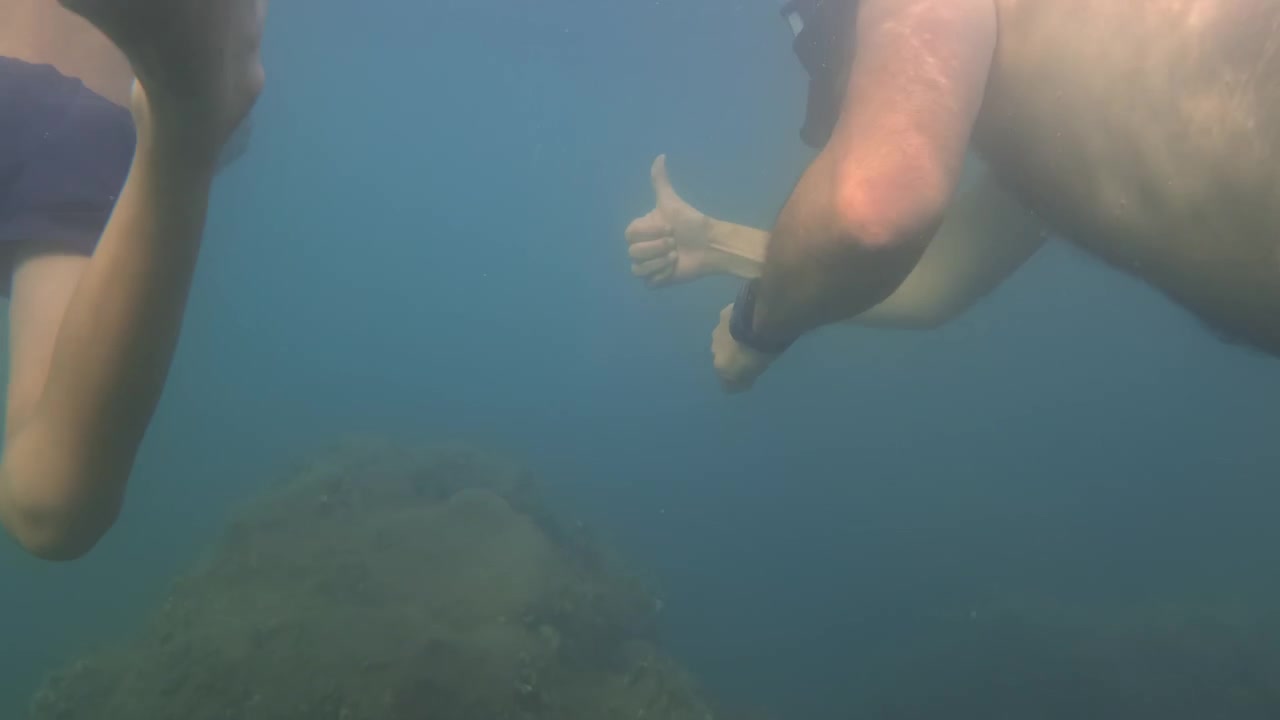 To personer, en mand og et barn, snorkler under vandet. De dykker nær klipper, udforsker havlivet og nyder sommerferien.