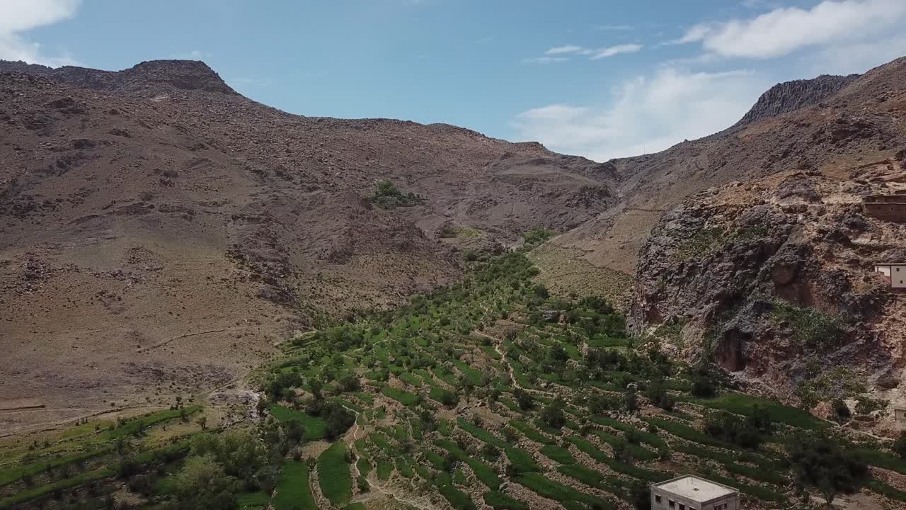 Aerial drone shot highlighting the contrast between arid geological rock faces and lush green terraced fields. Showcases natural erosion and mountain agriculture in the High Atlas
