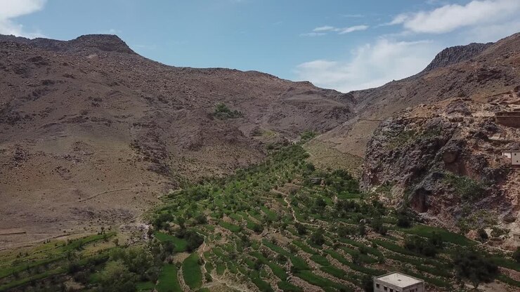 Aerial drone shot highlighting the contrast between arid geological rock faces and lush green terraced fields. Showcases natural erosion and mountain agriculture in the High Atlas
