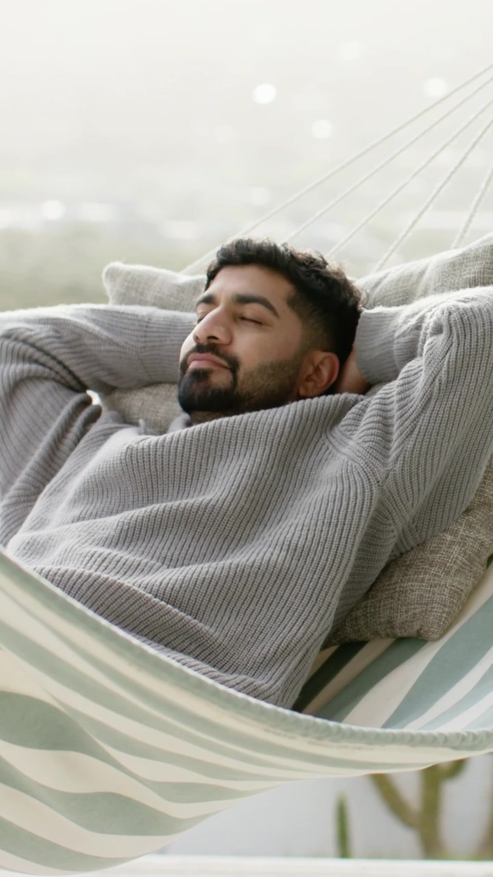 Video verticale: Uomo in maglia grigia sdraiato su amaca a righe sul balcone, mani dietro la testa.