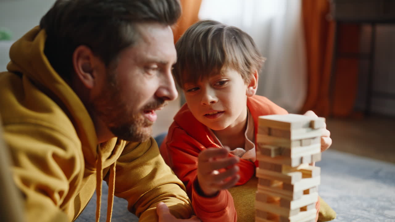 Padre che prende un blocco Jenga giocando con l'adorabile figlio, sdraiati sul pavimento della stanza, in primo piano.