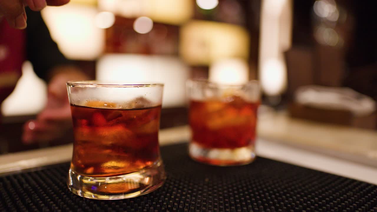 Bartender adds orange peel garnish to whiskey cocktail in dimly lit bar, close-up shot