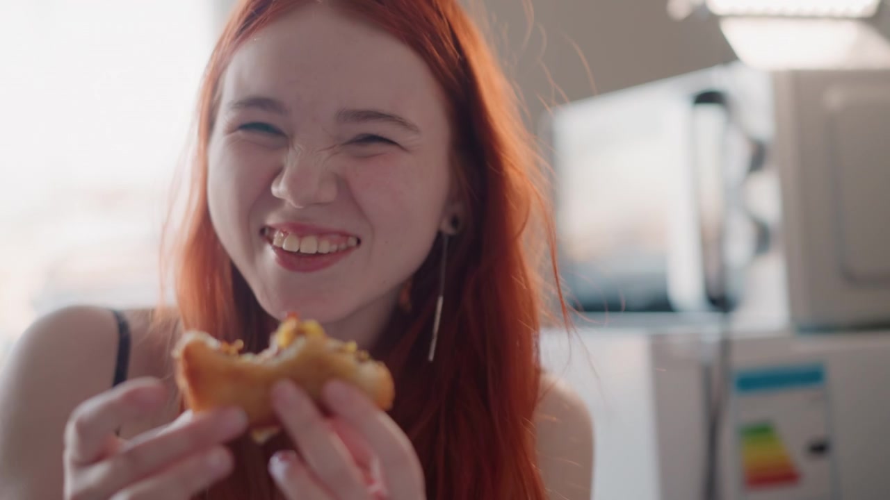 Ung kvinne spiser bakverk på kjøkkenet, lekent morgen-nærbilde viser smilende rødhåret som nyter flakete bakverk, mildt sollys, uformelt antrekk, avslappet uttrykk, fingre fulle av smuler, ærlig