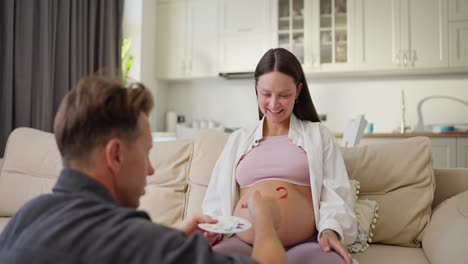 over his shoulder a man in a gray shirt paints with bright colors on the belly of his pregnant wife at home in a modern apartment