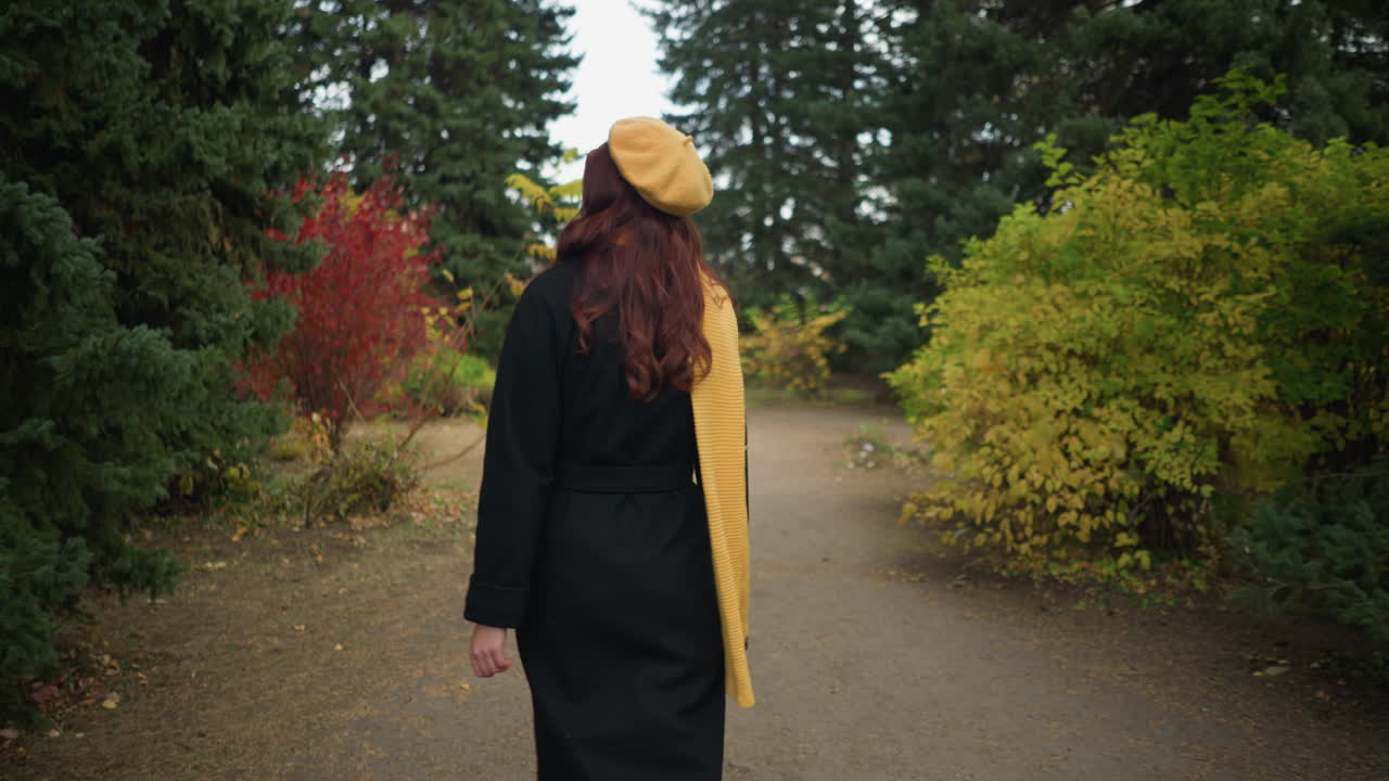 Vista posteriore di una giovane donna con lunghi capelli rossi ondulati, che indossa un cappotto nero, un berretto giallo e una sciarpa abbinata, mentre cammina lungo un sentiero sterrato attraverso un lussureggiante parco verde con un colorato fogliame autunnale.
