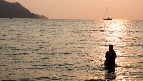 a woman fishing silhouetted against a beautiful sunset and yacht in the background on the horizon in bangsaray near pattaya, thailand