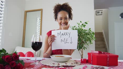 Mixed-race-woman-on-a-valentines-date-video-call,-holding-marry-me-proposal-sign