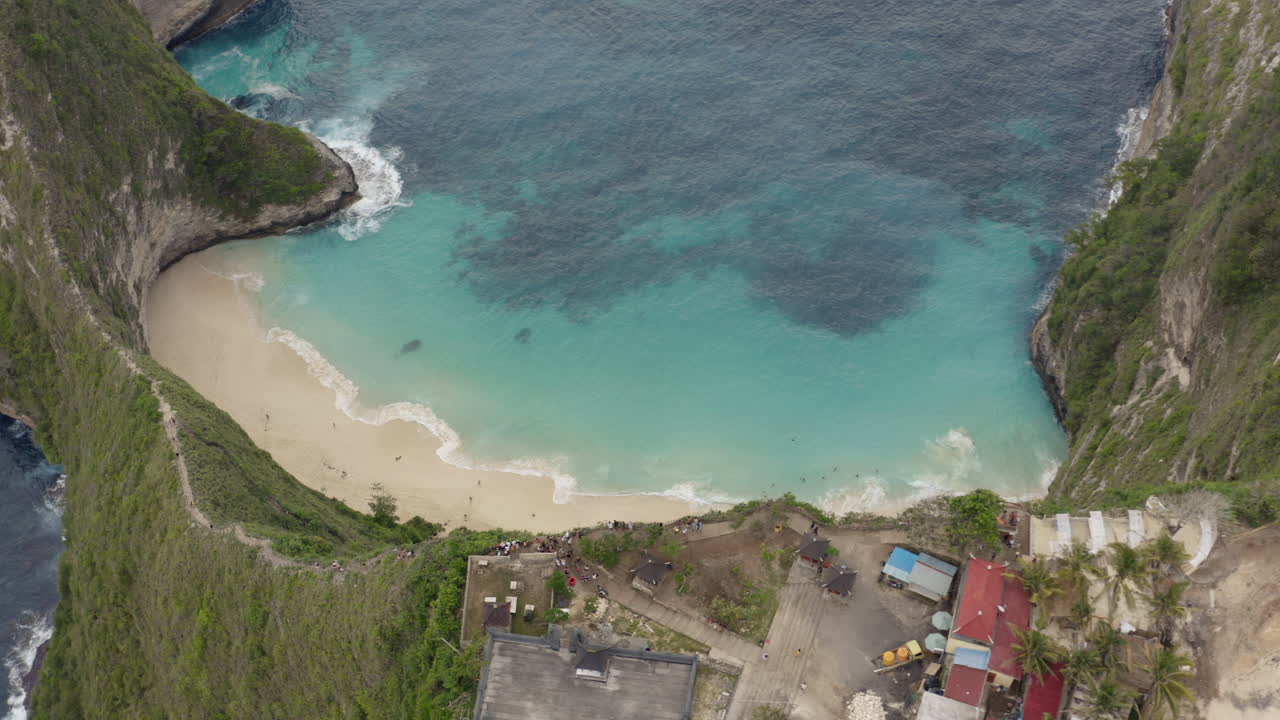 Slow pivot over Kelingking Beach surrounded by high steep cliffs with tourists walking on the sand and scenic spot viewpoint on top, Nusa Penida, Bali, Indonesia