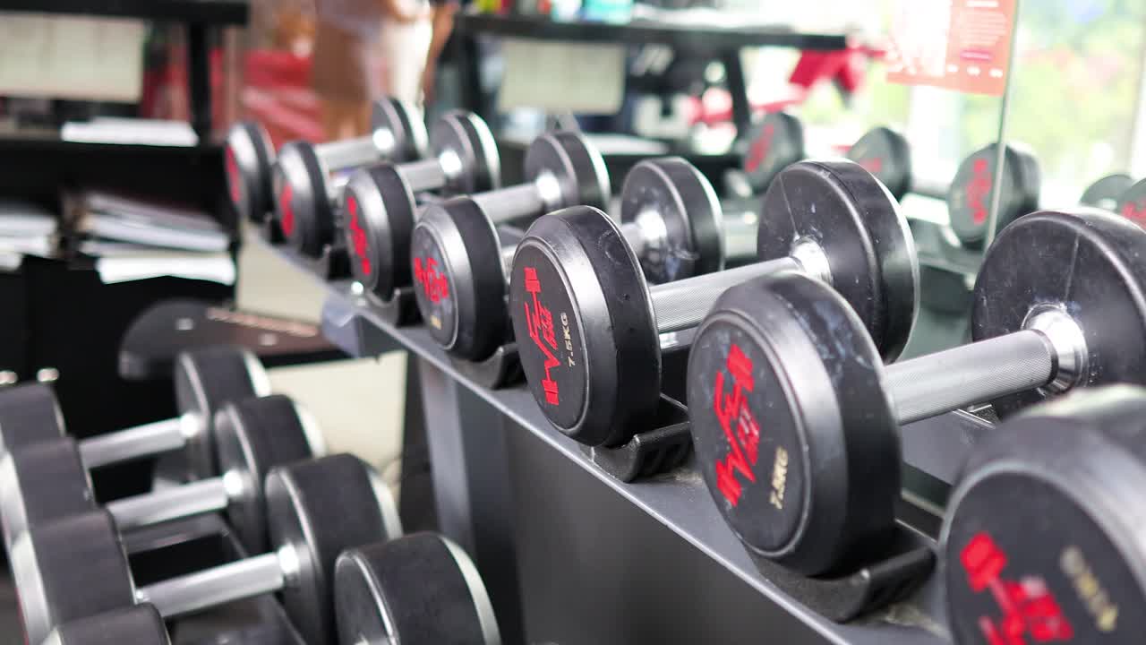 Rows of dumbbells in a well-lit gym, emphasizing fitness and strength training. The camera captures the equipment from various angles