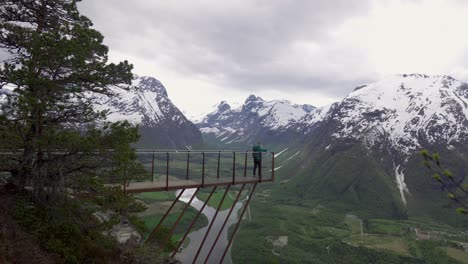 girl walking out to viewpoint on a metal bridge, watching over beautiful scenic view of snowy mountains, meadows and rivers in a valley