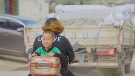 chinese boy smiles at camera as he rides on back of motorcycle driven by his mom in rural china.