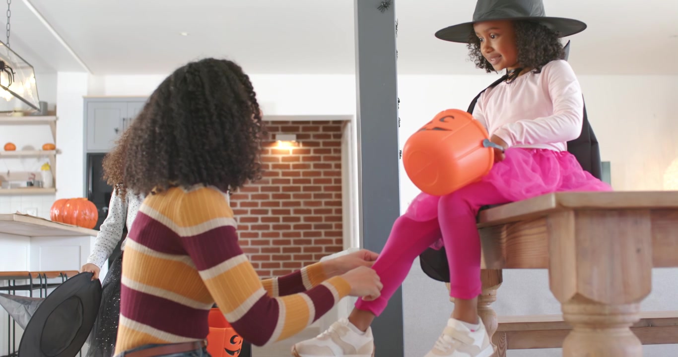 ハロウィンのため、自宅で娘がカボチャを持ちながら、アフリカ系アメリカ人の母親が娘の靴ひもを結んでいる様子
