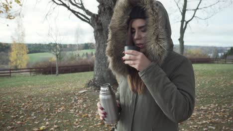 una chica adulta está sirviendo su bebida caliente y bebiéndola en un frío día de otoño