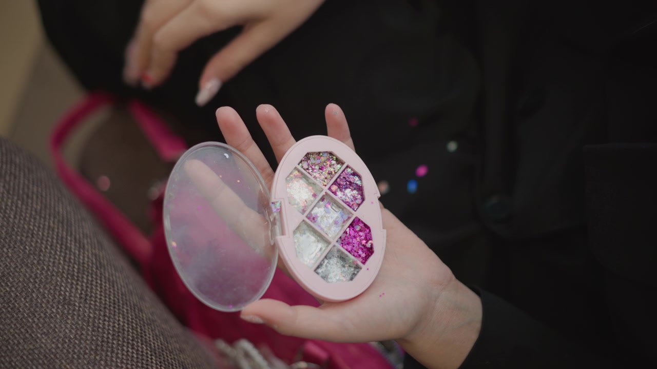 Mani di donna adulta tengono aperto un porta cipria rotondo con coperchio a specchio mentre le dita si librano sopra quattro cialde di ombretto scintillante nei toni del rosa e del viola, suggerendo selezione, prova e routine di bellezza.