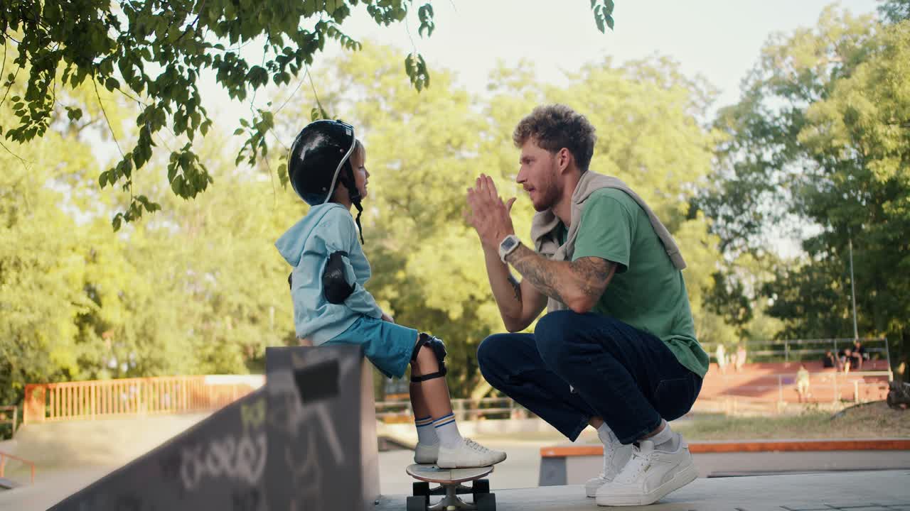 un niño pequeño con un suéter azul y un casco de seguridad negro y su padre con el cabello rizado en una camiseta verde se dan cinco en una patineta en el parque