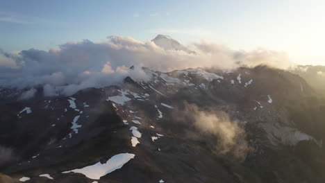 Drone-retreating-view-of-Mt-Baker-and-snow-pockets-on-ridgeline-with-yellow-clouds-at-sunset