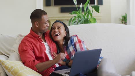 Happy-biracial-couple-sitting-on-sofa,-embracing,-using-laptop-and-laughing