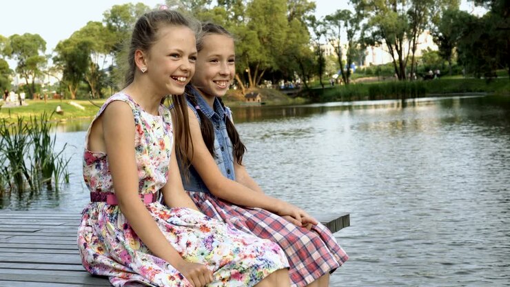 Duas meninas alegres sentam-se em uma ponte de madeira no verão, à beira do rio no Parque da cidade, uma ao lado da outra, olham para o lado e riem. Retrato.
