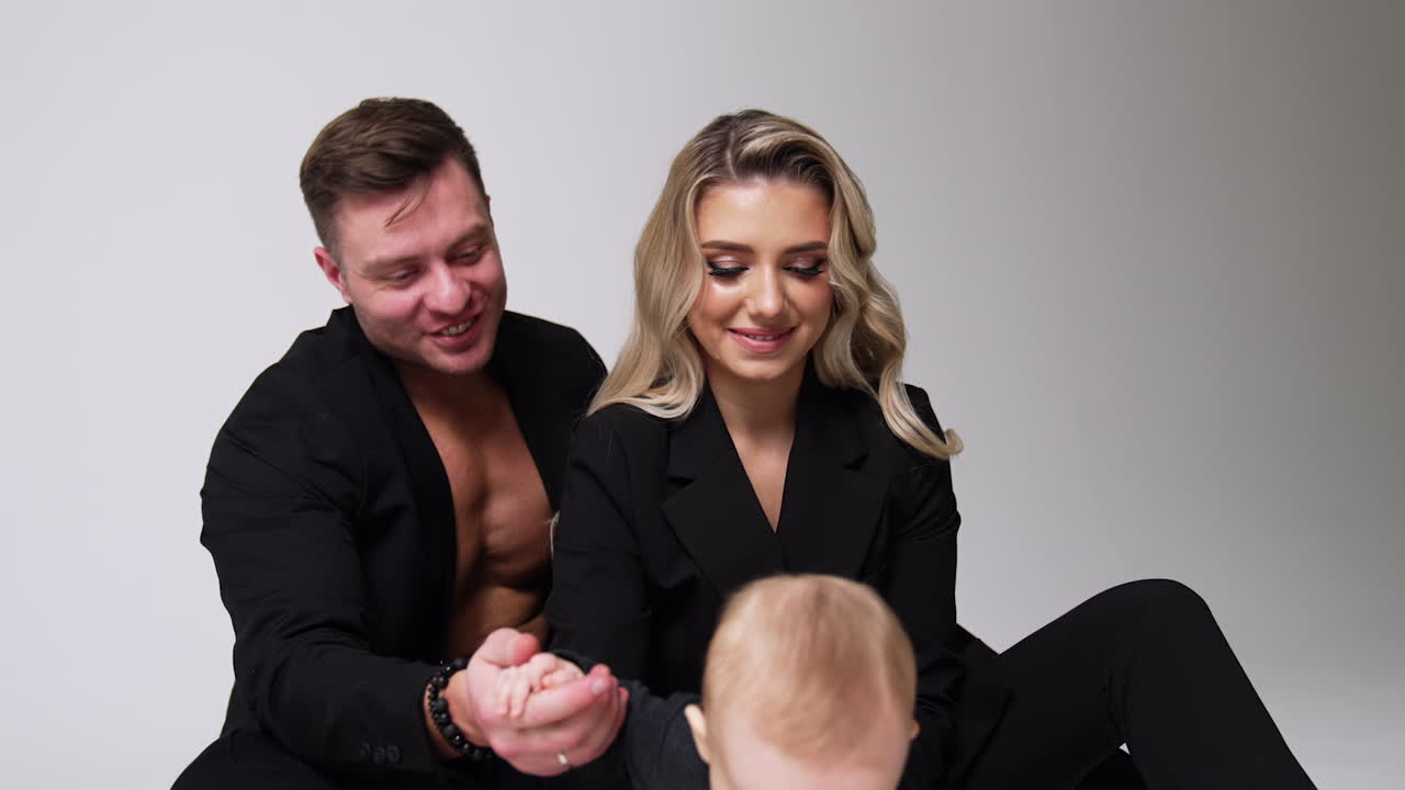 Beautiful Caucasian family of three. Couple wearing black suits and sitting on the floor supporting their little baby. White backdrop.
