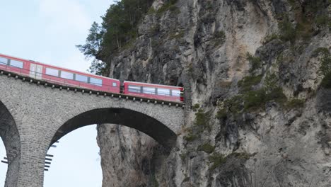 Traditional-red-Swiss-panorama-train-passing-on-a-stone-bridge-viaduct-into-a-mountain-tunnel-in-Landwasser-Brucke