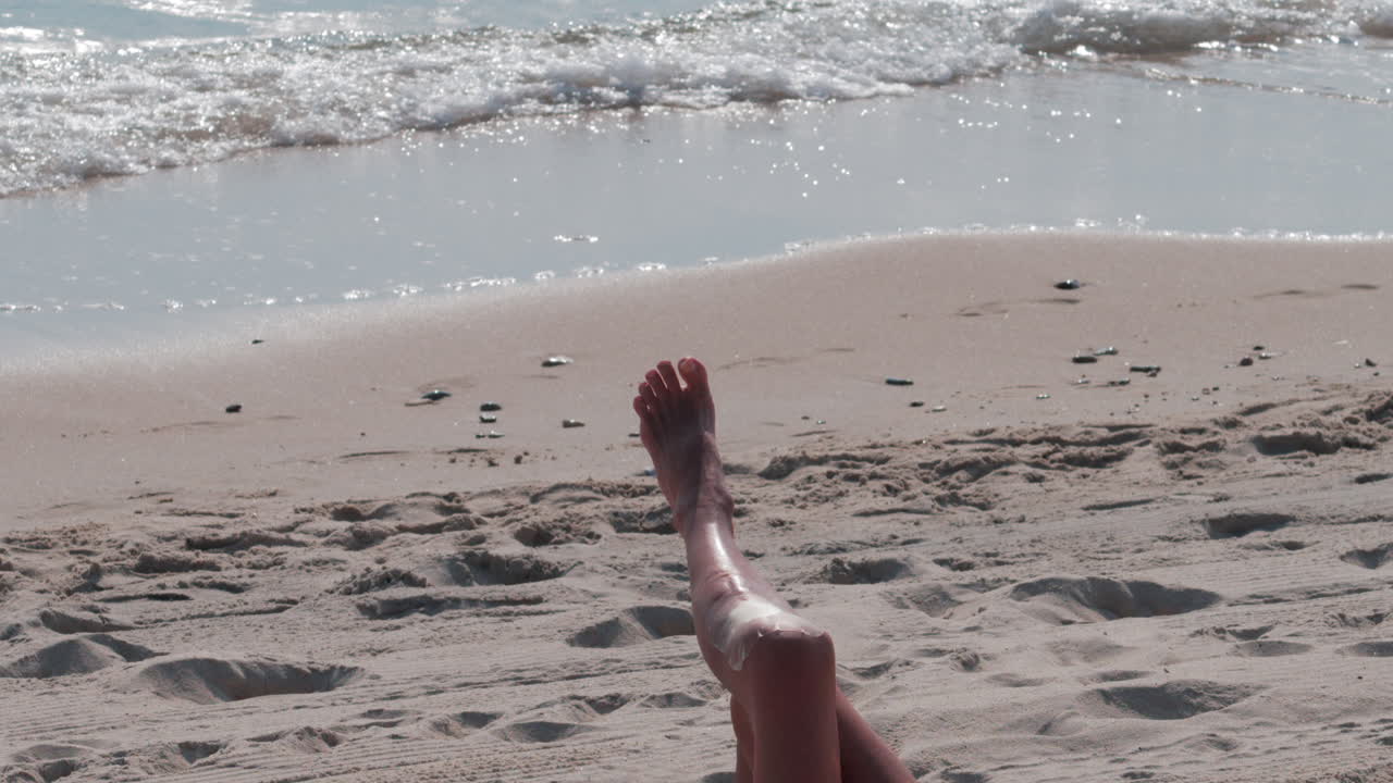 Close up of a woman's legs stretched on the beach as gentle waves roll in