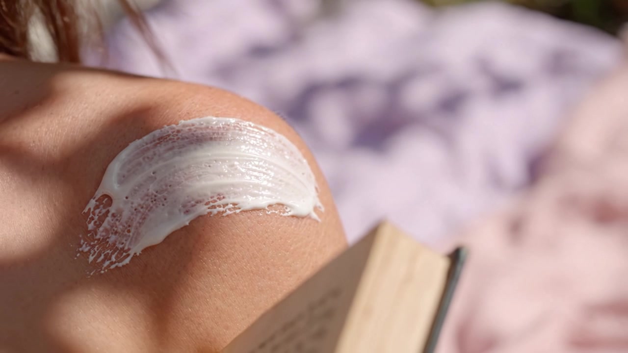 Applying Moisturizing Cream on Shoulder While Reading Book in the Sun, Close-up of Skincare Routine for Healthy Skin and Relaxation Outdoors