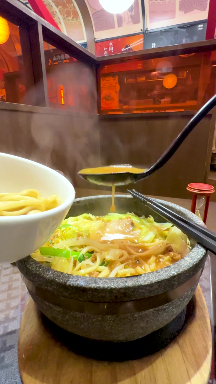 Eine Person gießt Brühe in eine dampfende Steinschale mit Ramen-Nudeln in einem Restaurant.