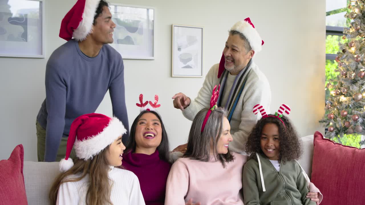 Diverse multi-generational family in Santa hats laughing after man playfully gesturing on red couch