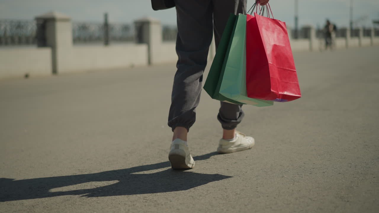 빨간색, 초록색 및 민트색 쇼핑 가방을 손에 들고 도로를 고 있는 사람의 다리 시각, 멀리서 자전거를 타고 있는 사람을 특징으로 하는 흐릿한 배경